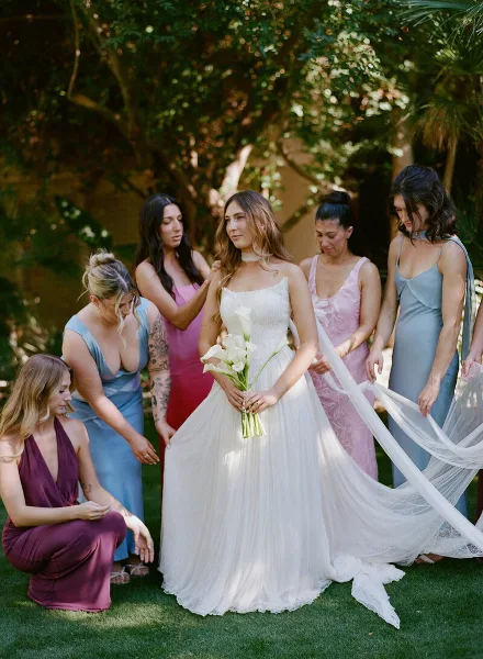 A bride in a white gown holding a bouquet of calla lilies, surrounded by bridesmaids in pastel dresses, standing outdoors in a garden.