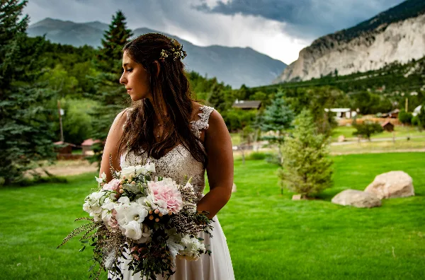 A bride in a white lace dress holding a bouquet of white and pink flowers, standing on a green lawn with mountains in the background.