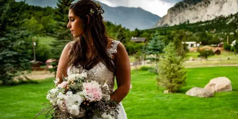 A bride in a white lace dress holding a bouquet of white and pink flowers, standing on a green lawn with mountains in the background.