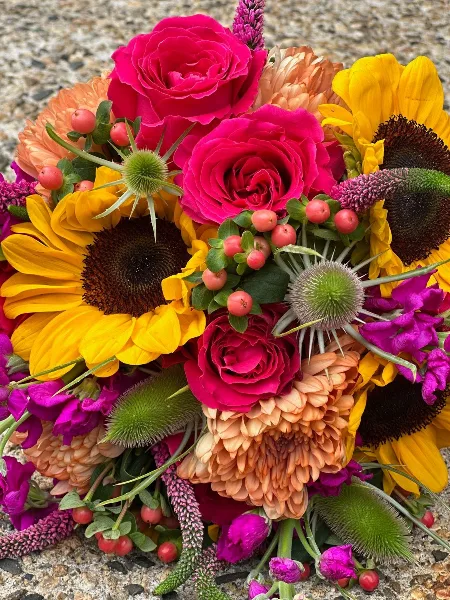 A close-up of a colorful wedding bouquet featuring bright yellow sunflowers, hot pink roses, orange dahlias, and green thistle buds.