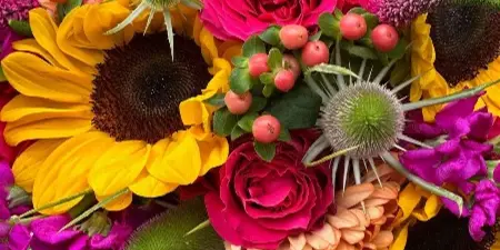 A close-up of a colorful wedding bouquet featuring bright yellow sunflowers, hot pink roses, orange dahlias, and green thistle buds.