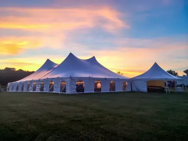 A large white event tent with multiple peaks set up on a grassy field at sunset.