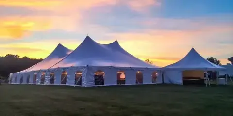 A large white event tent with multiple peaks set up on a grassy field at sunset.