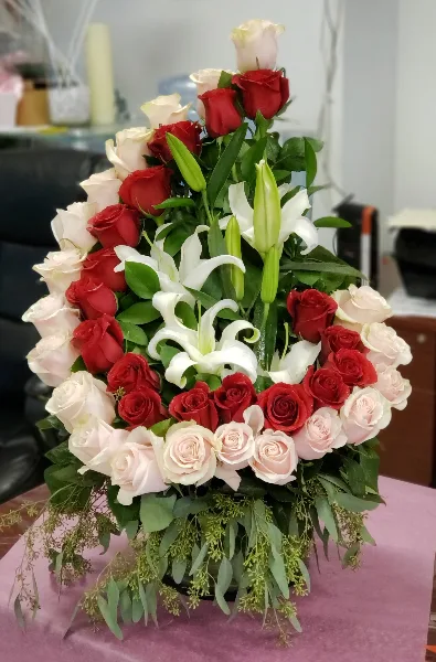 A tall floral arrangement featuring red, white, and blush roses with white lilies and greenery on a table.