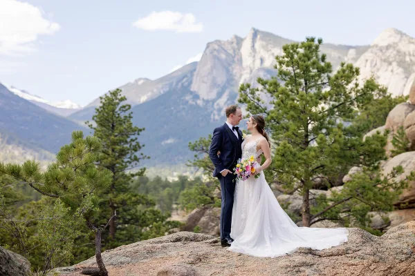 A bride and groom standing on a rocky outcrop with a mountain range in the background.