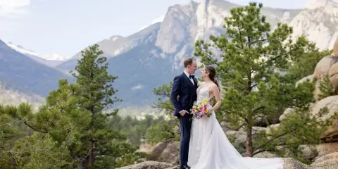 A bride and groom standing on a rocky outcrop with a mountain range in the background.