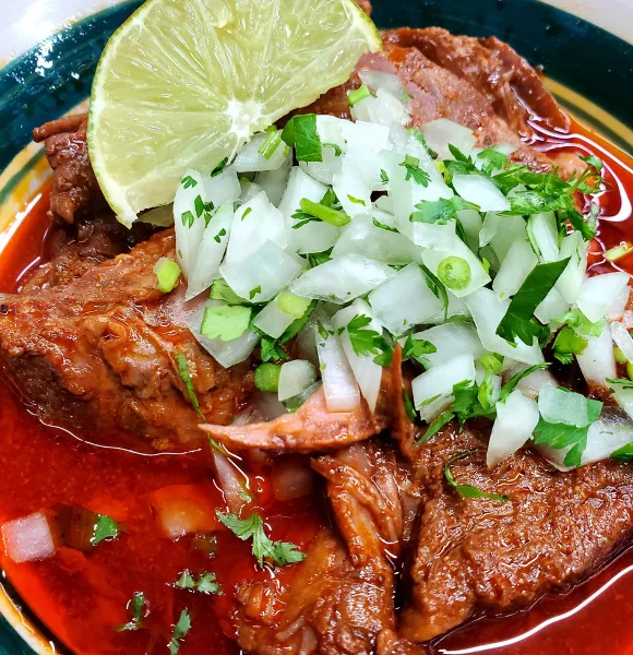 A plate of Mexican-style shredded meat in red sauce topped with diced white onion, cilantro, and a lime wedge.
