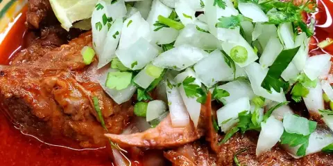 A plate of Mexican-style shredded meat in red sauce topped with diced white onion, cilantro, and a lime wedge.