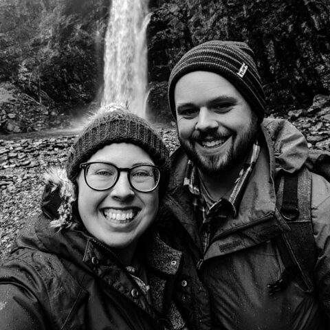 Black and white selfie of two smiling photographers wearing winter hats and jackets in front of a waterfall.