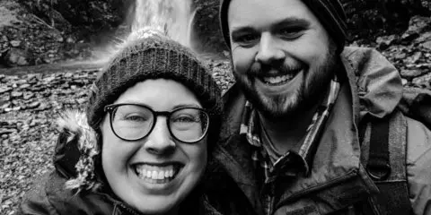 Black and white selfie of two smiling photographers wearing winter hats and jackets in front of a waterfall.