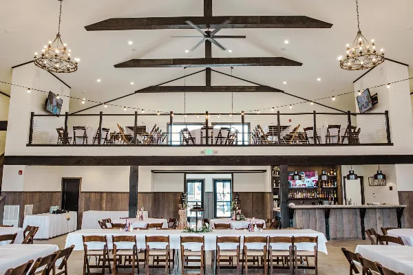 Interior view of a wedding venue hall with exposed wooden beams, chandeliers, and tables set with white linens.
