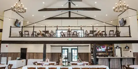 Interior view of a wedding venue hall with exposed wooden beams, chandeliers, and tables set with white linens.
