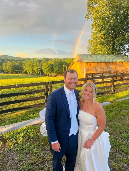 A bride and groom standing in front of a wooden farm pavilion with a rainbow in the sky and mountain views in the background.