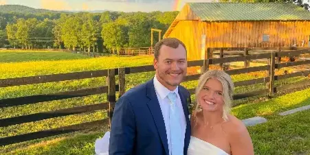 A bride and groom standing in front of a wooden farm pavilion with a rainbow in the sky and mountain views in the background.