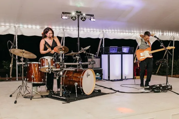 A male and female musician performing live on a stage with a drum set, electric guitar, and stage lighting.