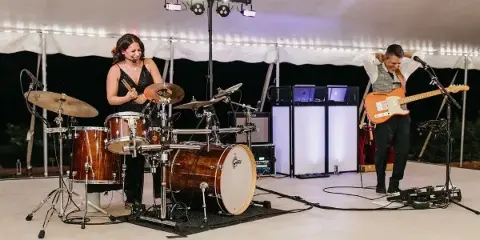 A male and female musician performing live on a stage with a drum set, electric guitar, and stage lighting.
