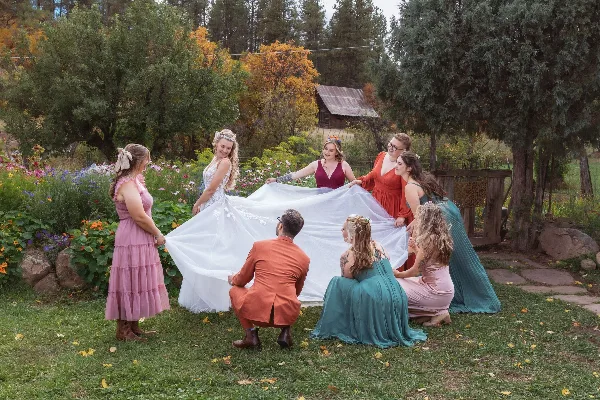 A bride in a white gown and a group of bridesmaids and a groom in colorful attire holding up the bride's train in a garden setting.