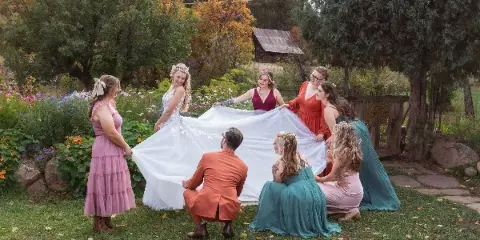 A bride in a white gown and a group of bridesmaids and a groom in colorful attire holding up the bride's train in a garden setting.