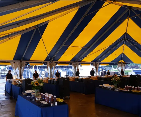 A catering event setup featuring a large blue and yellow striped tent, long buffet tables with food, and staff members in black shirts.