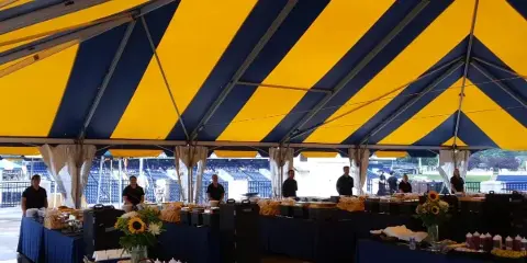 A catering event setup featuring a large blue and yellow striped tent, long buffet tables with food, and staff members in black shirts.