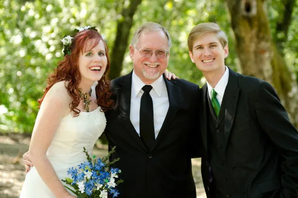 A smiling officiant in a black suit posing with a bride and groom outdoors.