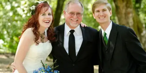 A smiling officiant in a black suit posing with a bride and groom outdoors.