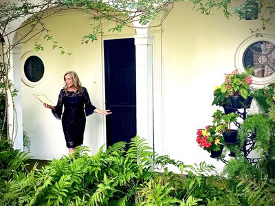 A woman in a black dress stands in a garden holding a book, representing a wedding officiant.
