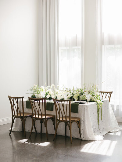 A styled wedding reception table with white floral arrangements, lace tablecloth, and wooden chairs in a bright room.