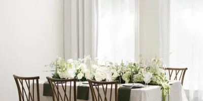 A styled wedding reception table with white floral arrangements, lace tablecloth, and wooden chairs in a bright room.