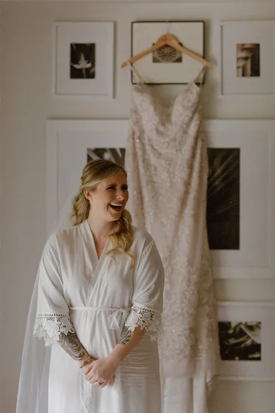 A bride in a white robe and veil laughing while standing in front of a hanging wedding dress.