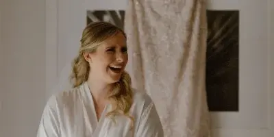 A bride in a white robe and veil laughing while standing in front of a hanging wedding dress.
