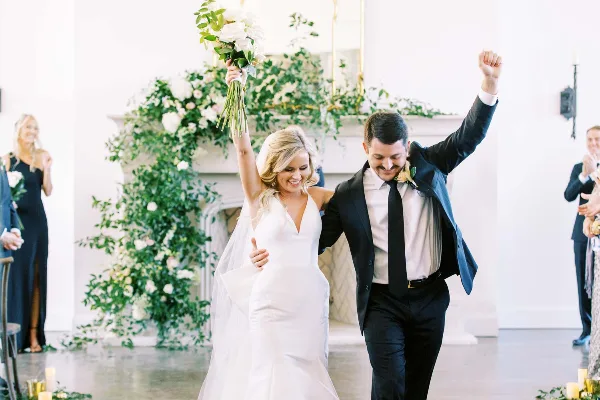 A bride and groom walking down the aisle with raised arms, smiling, in a bright white wedding dress and black suit.