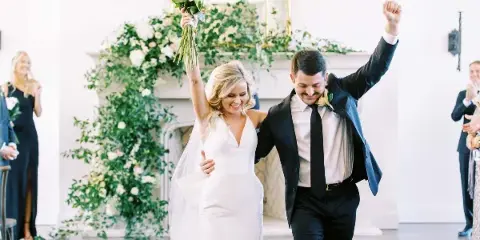 A bride and groom walking down the aisle with raised arms, smiling, in a bright white wedding dress and black suit.