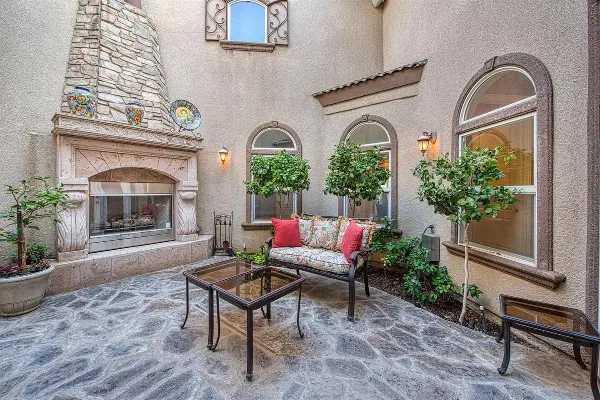 Outdoor courtyard of a Tuscan-style estate with a stone fireplace, garden trees, and a cushioned bench.