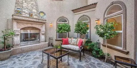 Outdoor courtyard of a Tuscan-style estate with a stone fireplace, garden trees, and a cushioned bench.