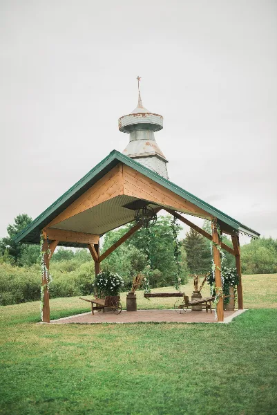 A wooden outdoor wedding pavilion with a green roof and cupola, decorated with hanging floral garlands and rustic wooden furniture on a brick patio.