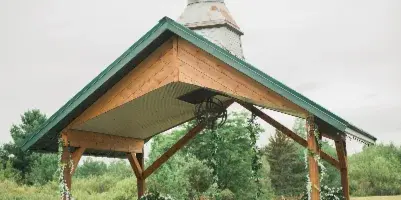 A wooden outdoor wedding pavilion with a green roof and cupola, decorated with hanging floral garlands and rustic wooden furniture on a brick patio.