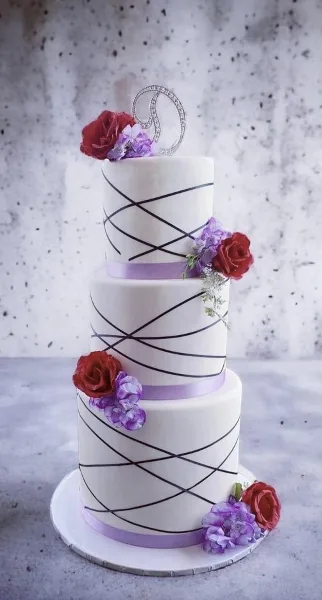 A three-tiered white wedding cake featuring black crisscross piping, purple ribbon accents, and red and purple sugar flowers.