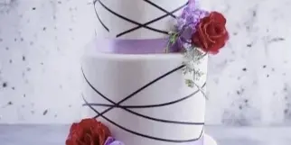 A three-tiered white wedding cake featuring black crisscross piping, purple ribbon accents, and red and purple sugar flowers.