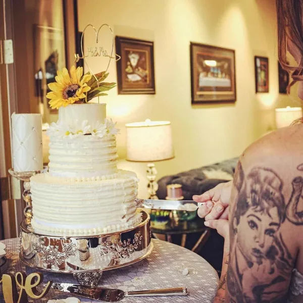 A three-tier white wedding cake with textured frosting, decorated with a sunflower and white daisies, sitting on a silver stand while a person cuts a slice.