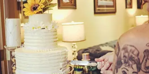 A three-tier white wedding cake with textured frosting, decorated with a sunflower and white daisies, sitting on a silver stand while a person cuts a slice.