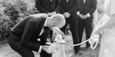 A black and white photograph of a groom kissing a dog during a wedding ceremony, with the bride and groomsmen looking on.