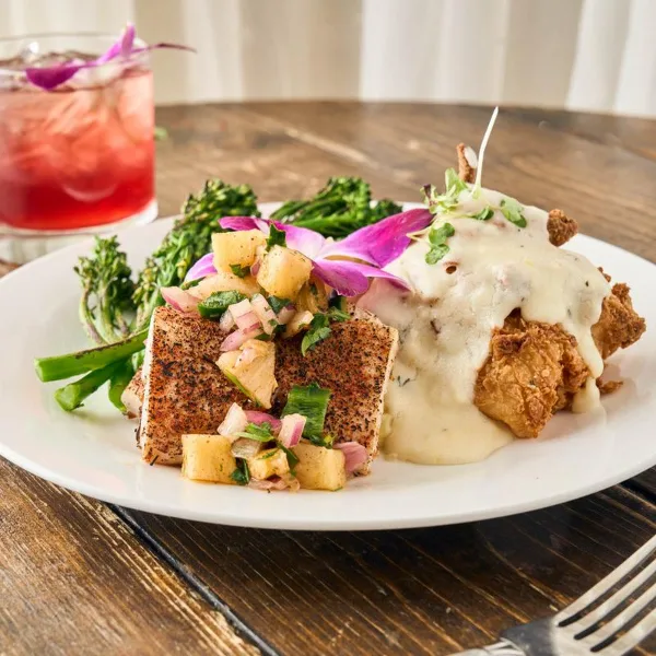 A plated meal featuring blackened fish fillet and fried chicken smothered in gravy, garnished with fresh vegetables and a flower.