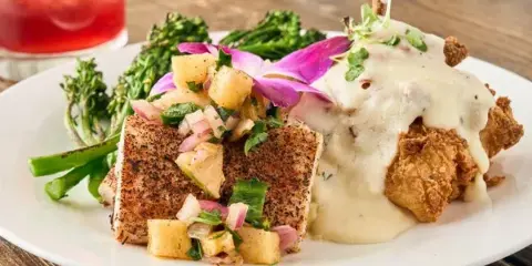 A plated meal featuring blackened fish fillet and fried chicken smothered in gravy, garnished with fresh vegetables and a flower.