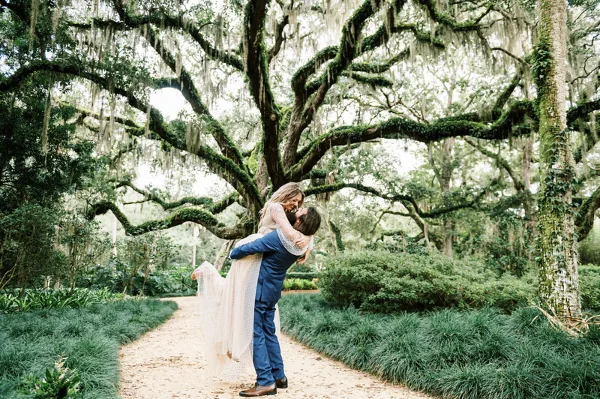 A bride and groom sharing a kiss on a path in a lush, moss-draped garden setting.