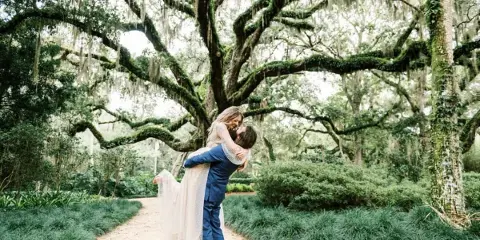 A bride and groom sharing a kiss on a path in a lush, moss-draped garden setting.