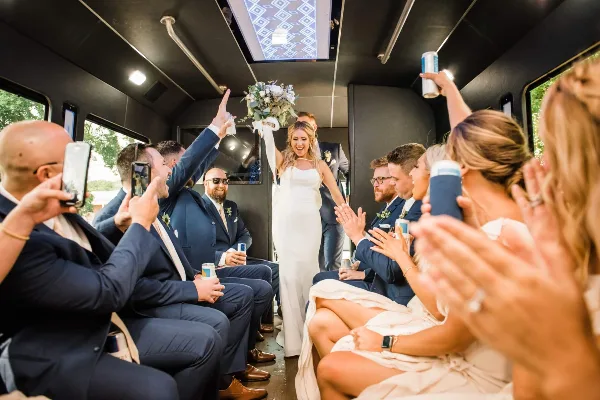 A bride stands in the aisle of a shuttle bus surrounded by a cheering wedding party holding drinks.