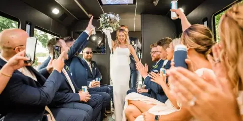 A bride stands in the aisle of a shuttle bus surrounded by a cheering wedding party holding drinks.