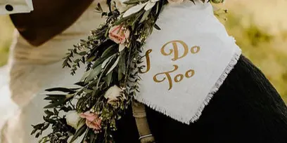 A black dog wearing a white bandana with 'I Do Too' text and a floral arrangement with pink roses and greenery.