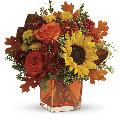 Autumn floral arrangement with red roses, sunflowers, and fall leaves in a square glass vase